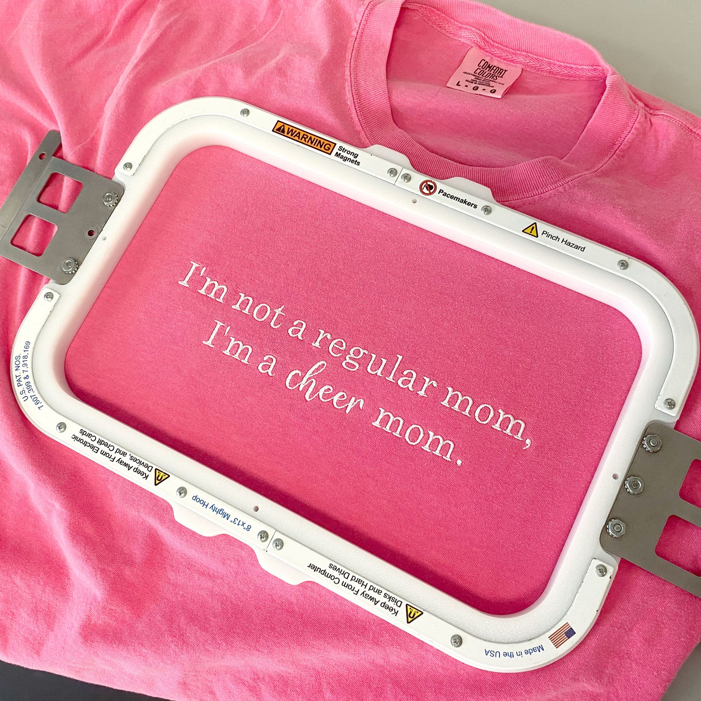 flat lay of a crucnhberry pink comfort colors tshirt with with Embroidered  I&#39;m not a regular mom, I'm a cheer mom design. shown in the embroidery hoop