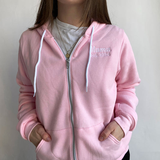 young woman wearing a pink bella and canvas full zip jacket with embroidered Mama est 2024 design on the left chest in white thread