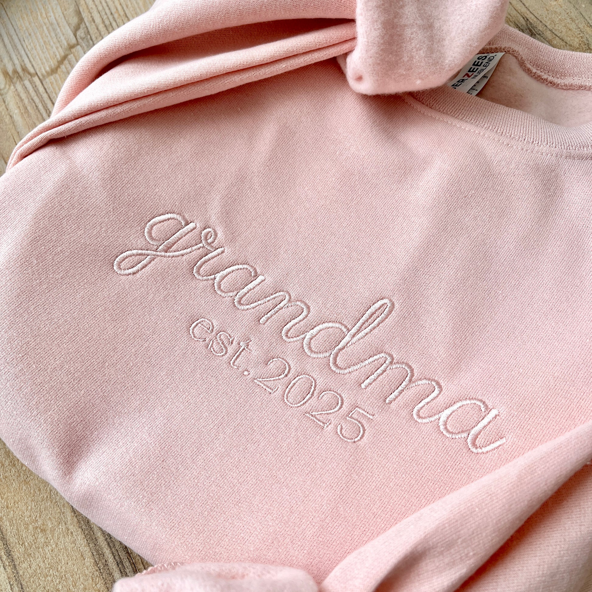 closeup of blush crewneck sweatshirt with custom embroidered grandma est. 2025 design in powder pink thread