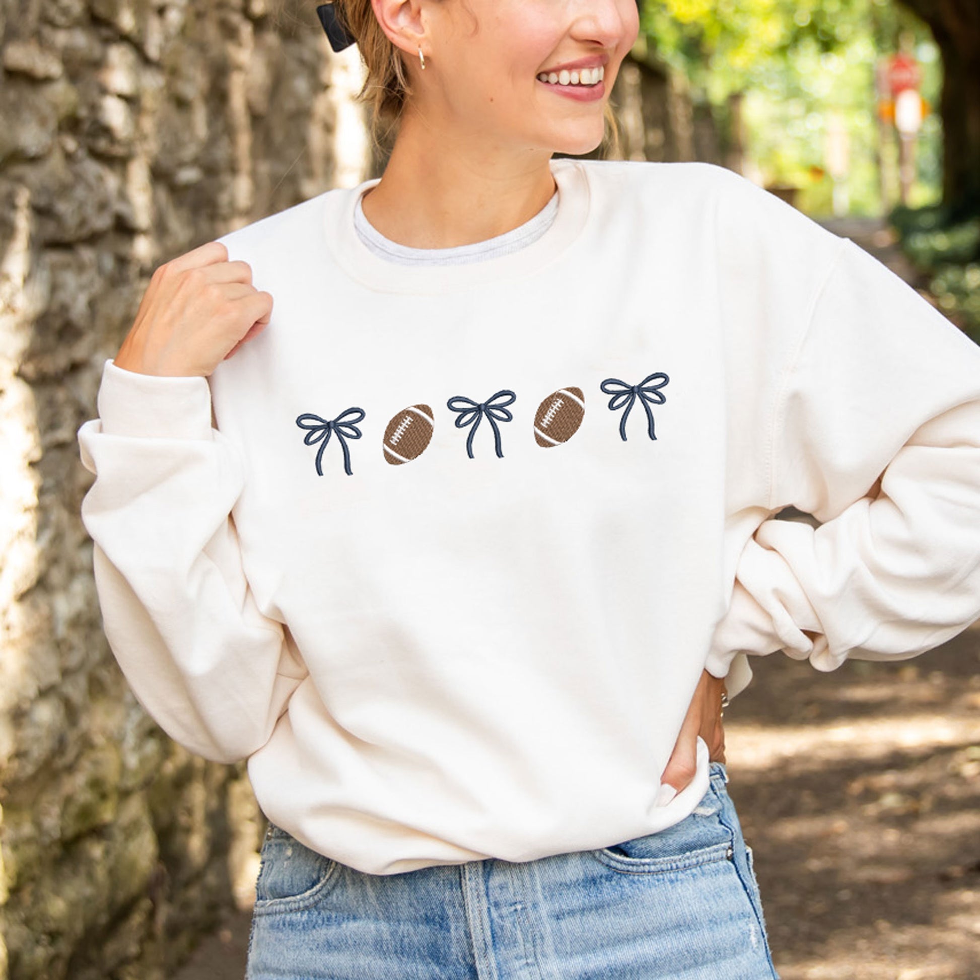 woman wearing a sweet cream crewneck sweatshirt with embroidered alternating bows and footballs design across the chest. The bow are embroidered in navy thread.