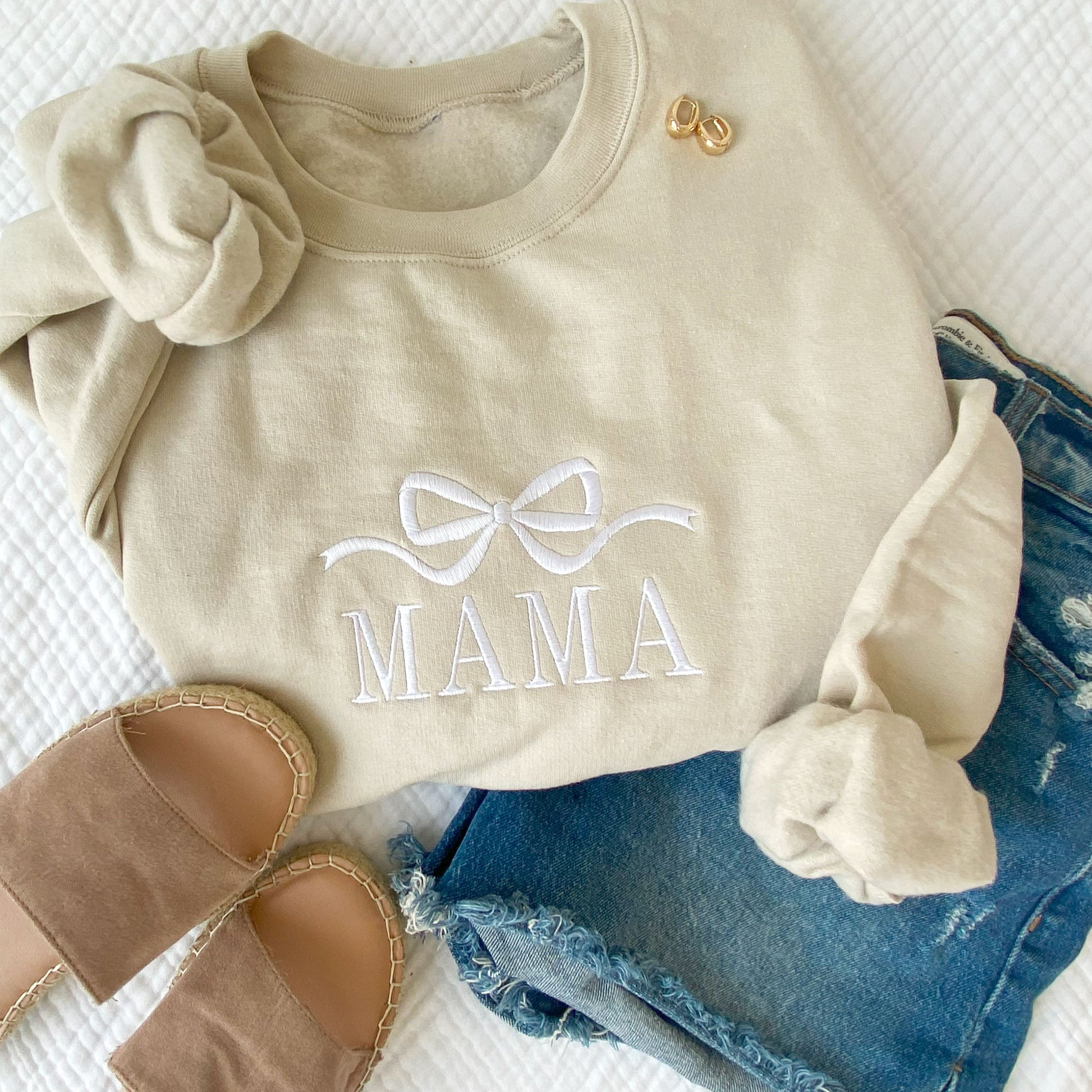 styled flat lay of a sand crewneck sweatshirt with jeans and sandals. On the crewneck is embroidered Bow and mama design in white thread
