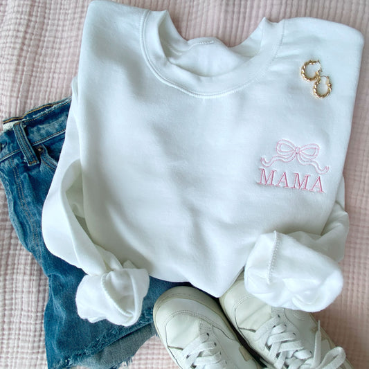 white crewneck sweatshirt with embroidered outline bow and all caps mama design on the left chest in baby pink thread styled with jean shorts and  sneakers