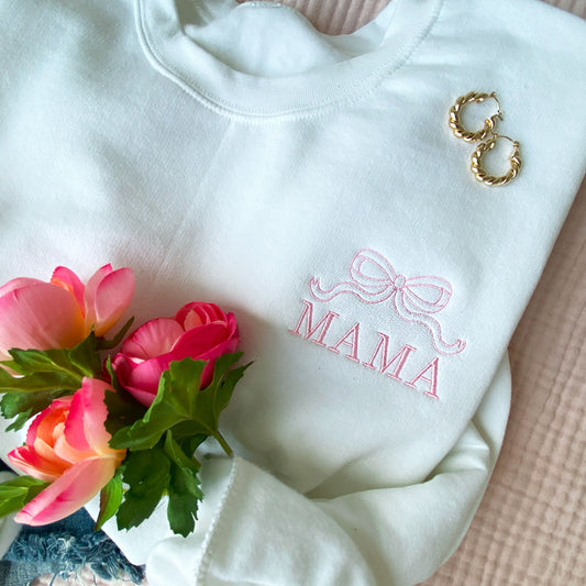 white crewneck sweatshirt with embroidered outline bow and all caps mama design on the left chest in baby pink thread styled with flowers and earrings