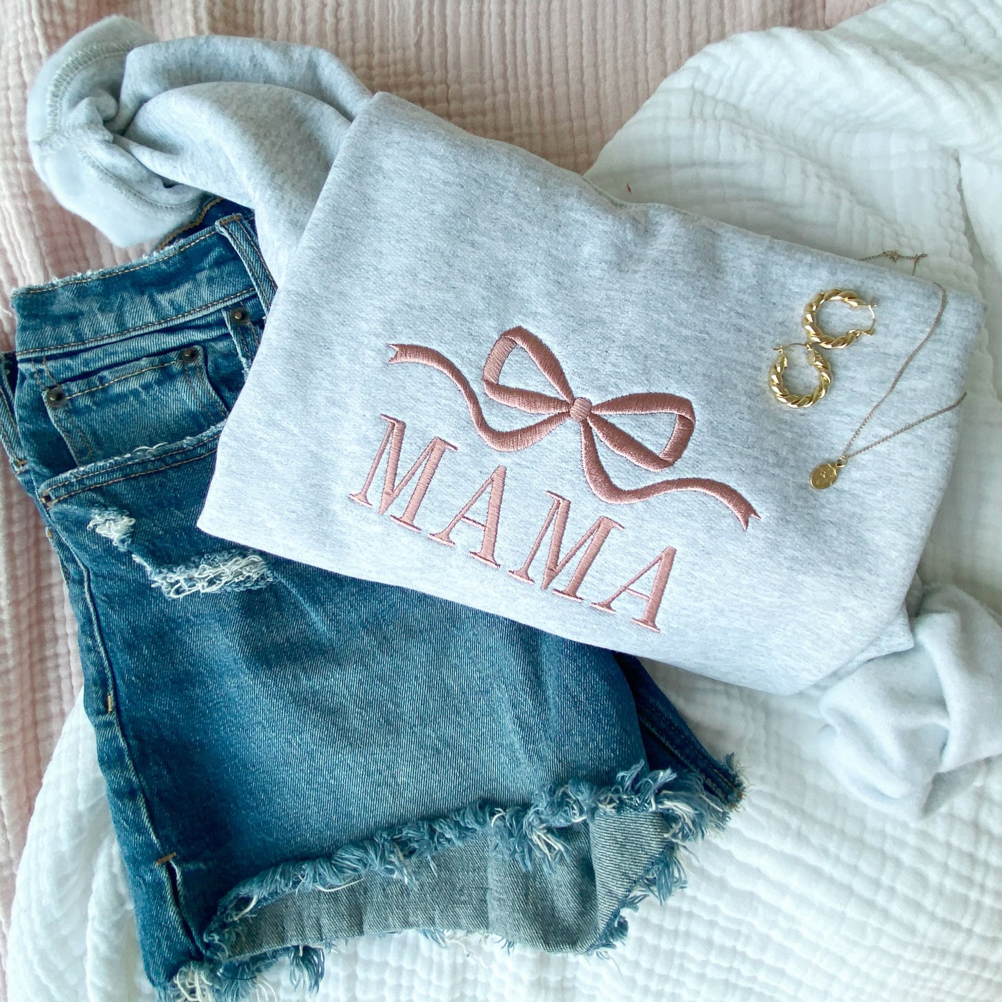 styled flat lay of an ash crewneck sweatshrt with jeans and jewelry. On the crewneck is embroidered Bow and mama design in mauve thread