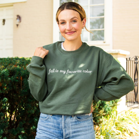 woman wearing jeans and a military green crewneck sweatshirt with a custom embroidery reading 'fall is my favorite color' in a natural thread color, standing outside a large cream brick house.