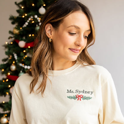 teacher wearing an ivory long sleeve comfort colors shirt with custom name and garland bow embroidery design on left chest