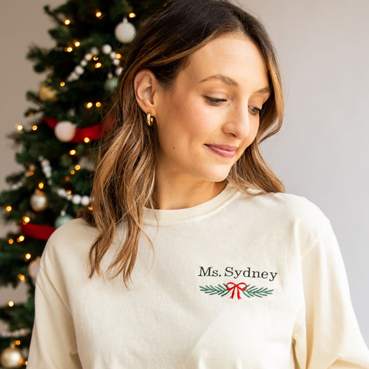 teacher wearing an ivory long sleeve comfort colors shirt with custom name and garland bow embroidery design on left chest