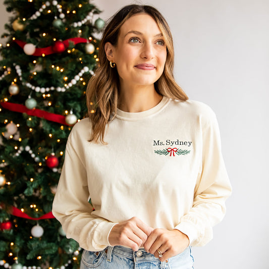teacher standing near christmas tree with a custom name and garland embroidered comfort colors long sleeve top