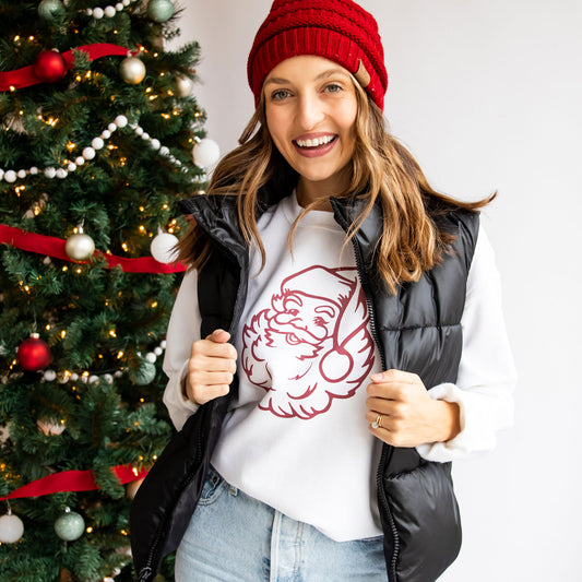 woman wearing a black puffer vest and red hat and a white crewneck sweatshirt with a red jolly santa print on the front