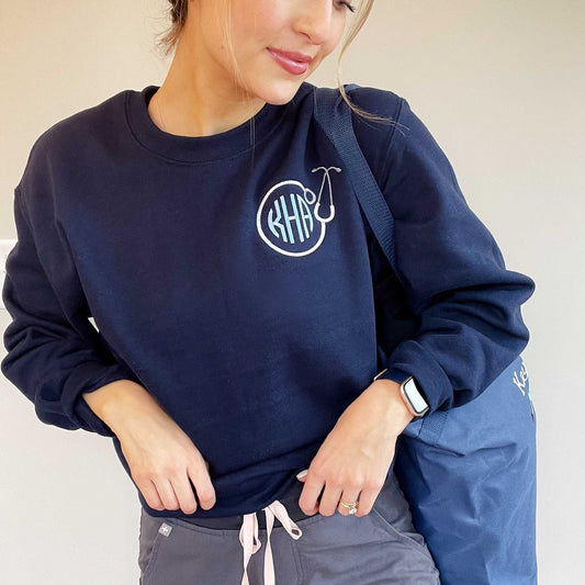nurse holding a tote bag and wearing gray scrub joggers, and a navy crewneck sweatshirt with a personalized round stethoscope and monogram embroidery design on the left chest
