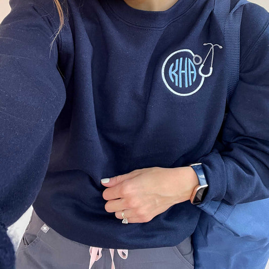 close up of nurse wearing a navy pullover sweatshirt with a custom stethoscope and monogram designed embroidered on the left chest