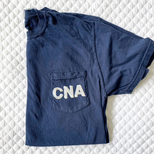dark blue tee with nursing credentials embroidered on the pocket in a shadow block font