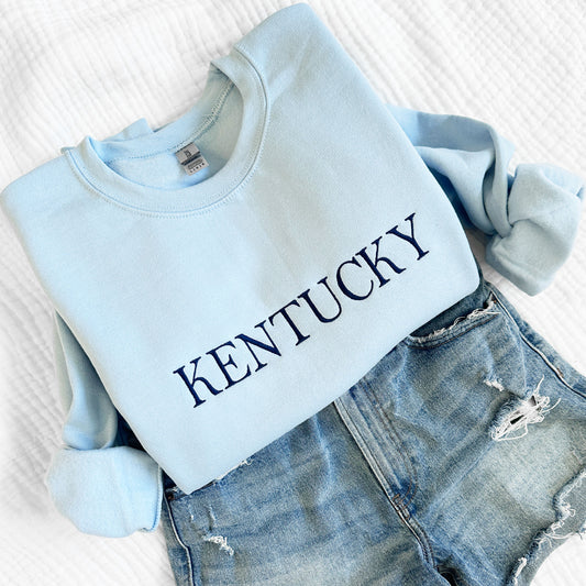 styled flat lay of a light blue crewneck sweatshirt with embroidered Kentucky in navy thread and shown with a pair of jean shorts