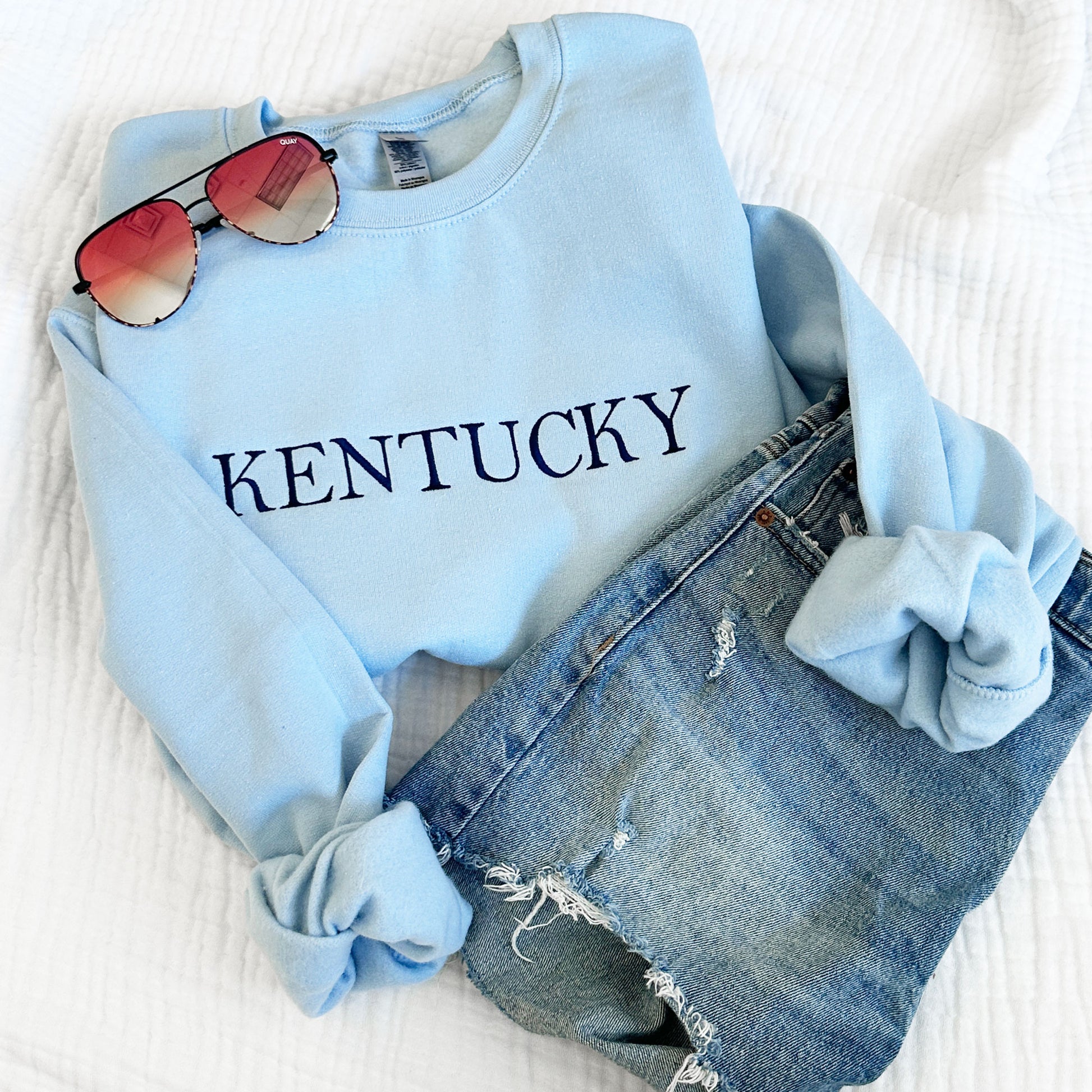 light blue crewneck sweatshirt with embroidered Kentucky in navy thread styled with jeans an sunglasses
