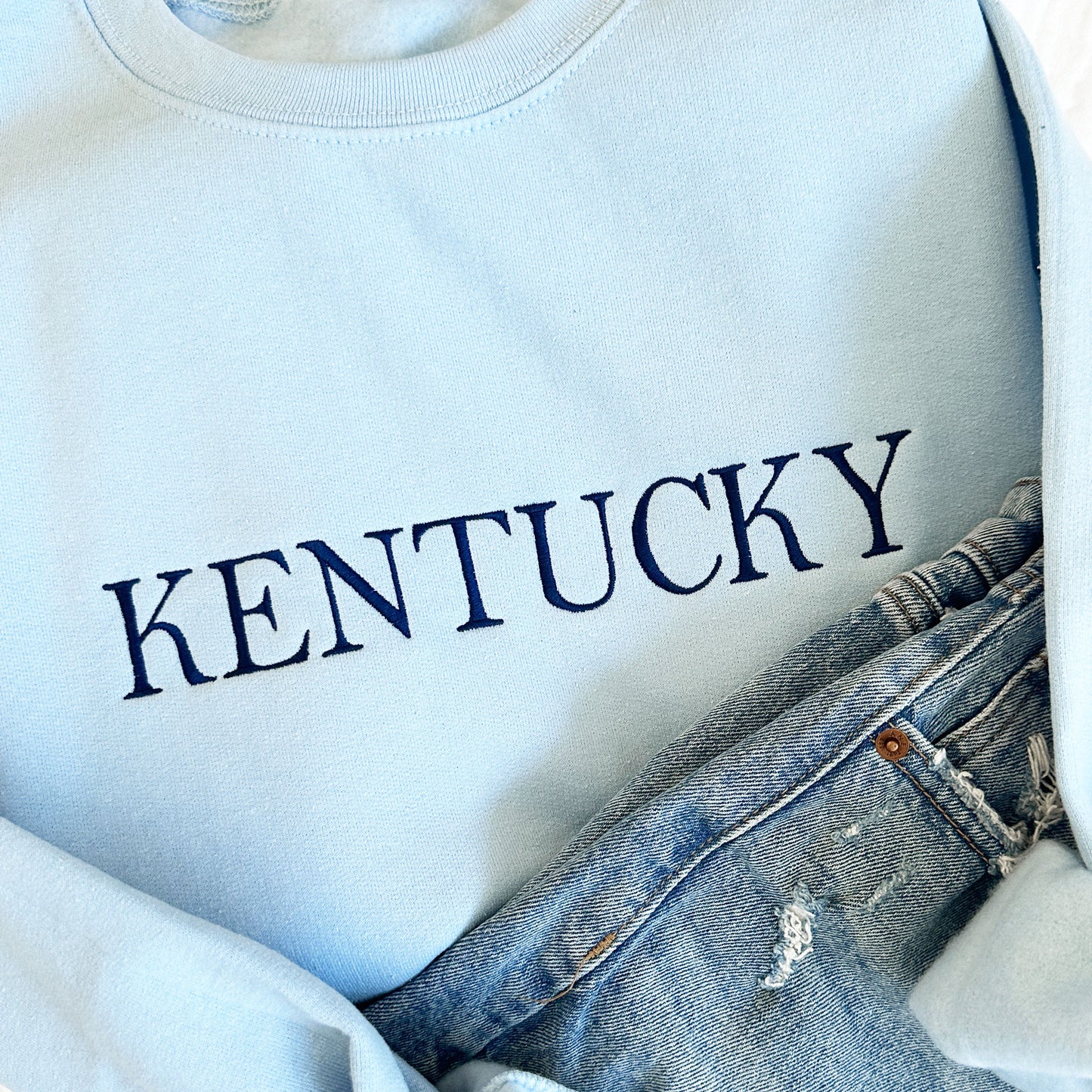 close up of a light blue crewneck sweatshirt with embroidered Kentucky in navy thread