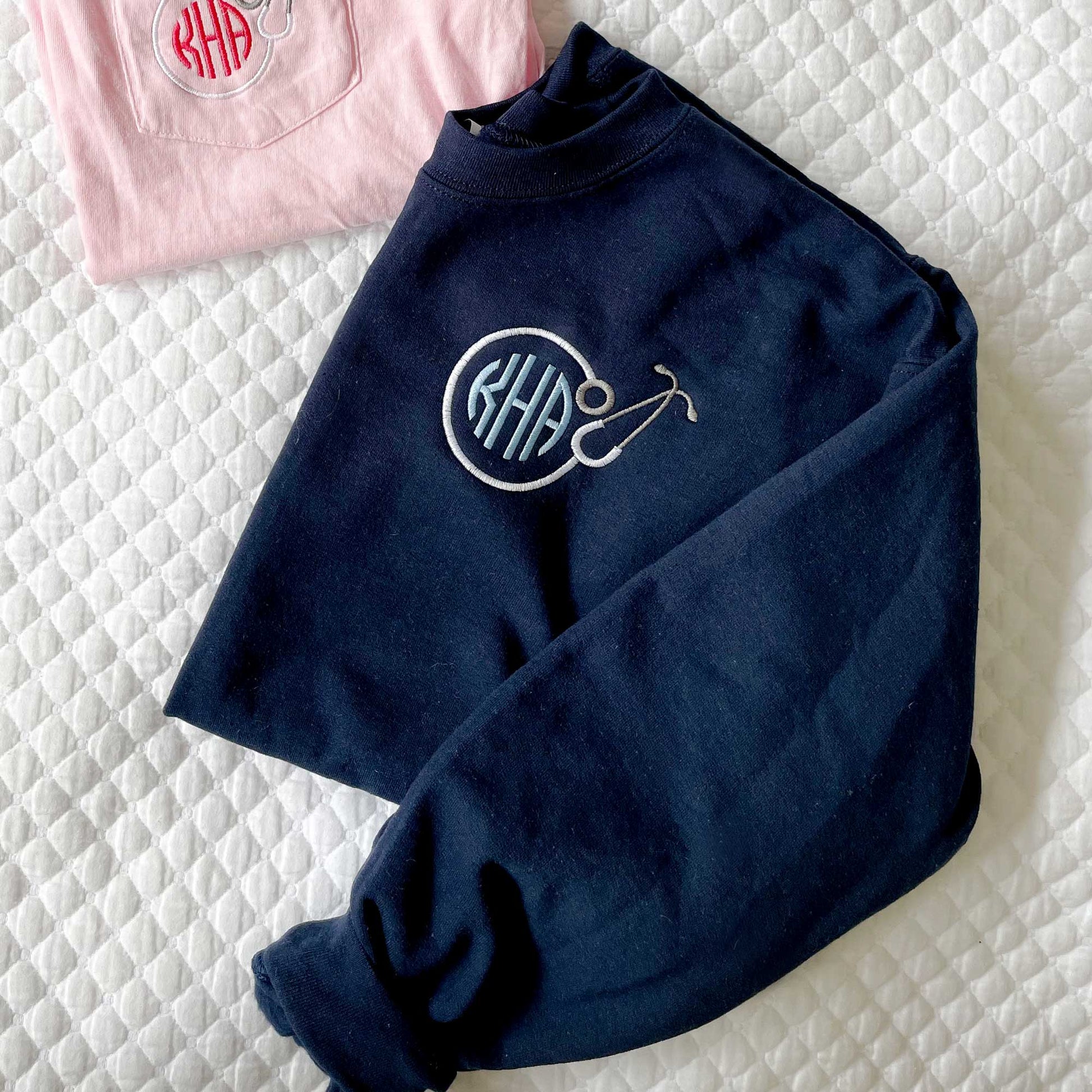flat lay photo of a navy crewneck sweatshirt with a personalized embroidery design on the left chest featuring a white ground stethoscope and a rounded baby blue three letter monogram inside of it.