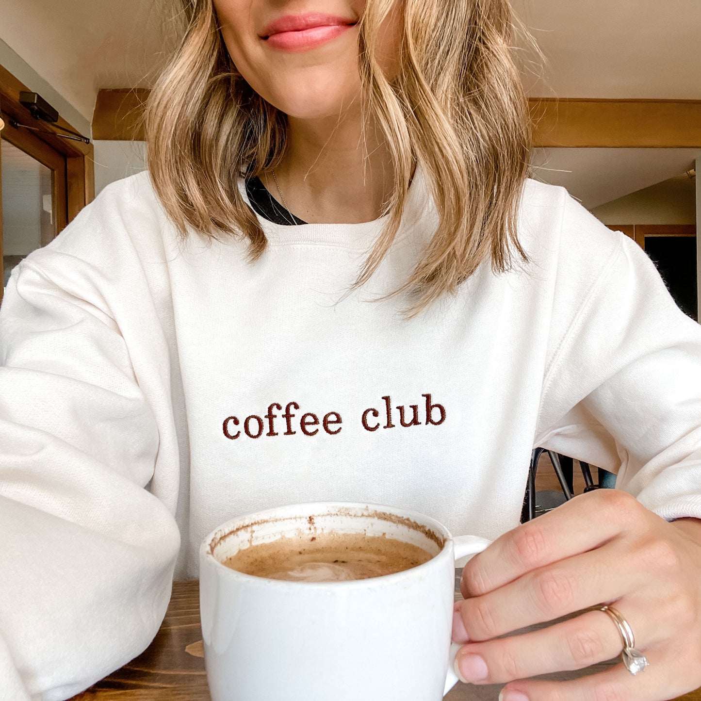 woman in a coffee shop wearing a sweet cream crewneck sweatshirt with embroidered coffee club across the chest in chestnut  thread