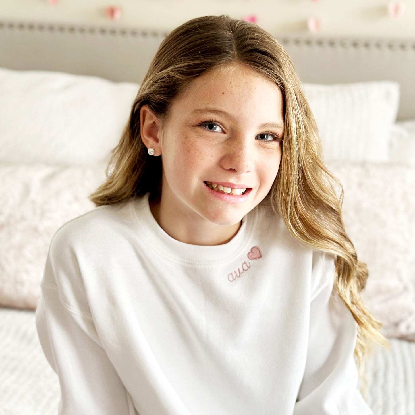 young girl wearing a white crewneck sweatshirt with her name ava and a heart embroidered in mauve along the neckline