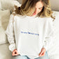 woman sitting on bed wearing a white sweatshirt with an in my lover era embroidered design on the front