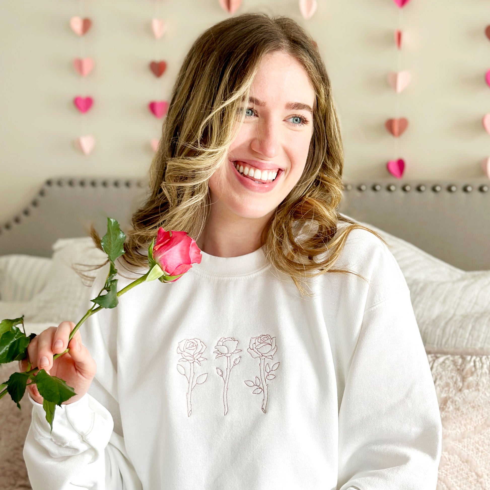 young woman holding a rose and modeling a white crewneck sweatshirt with three embroidered roses across the chest