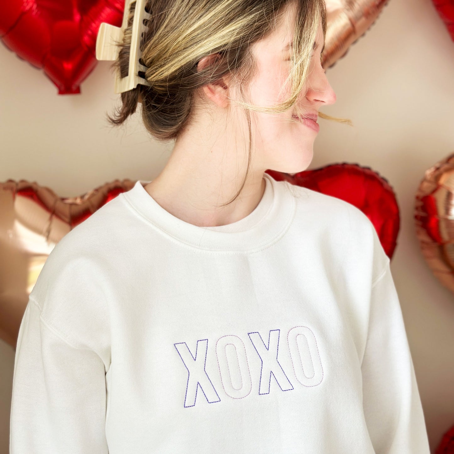 young woman wearing a white crewneck sweatshirt that features purple and lilac stitched xoxo design embroidered across the chest