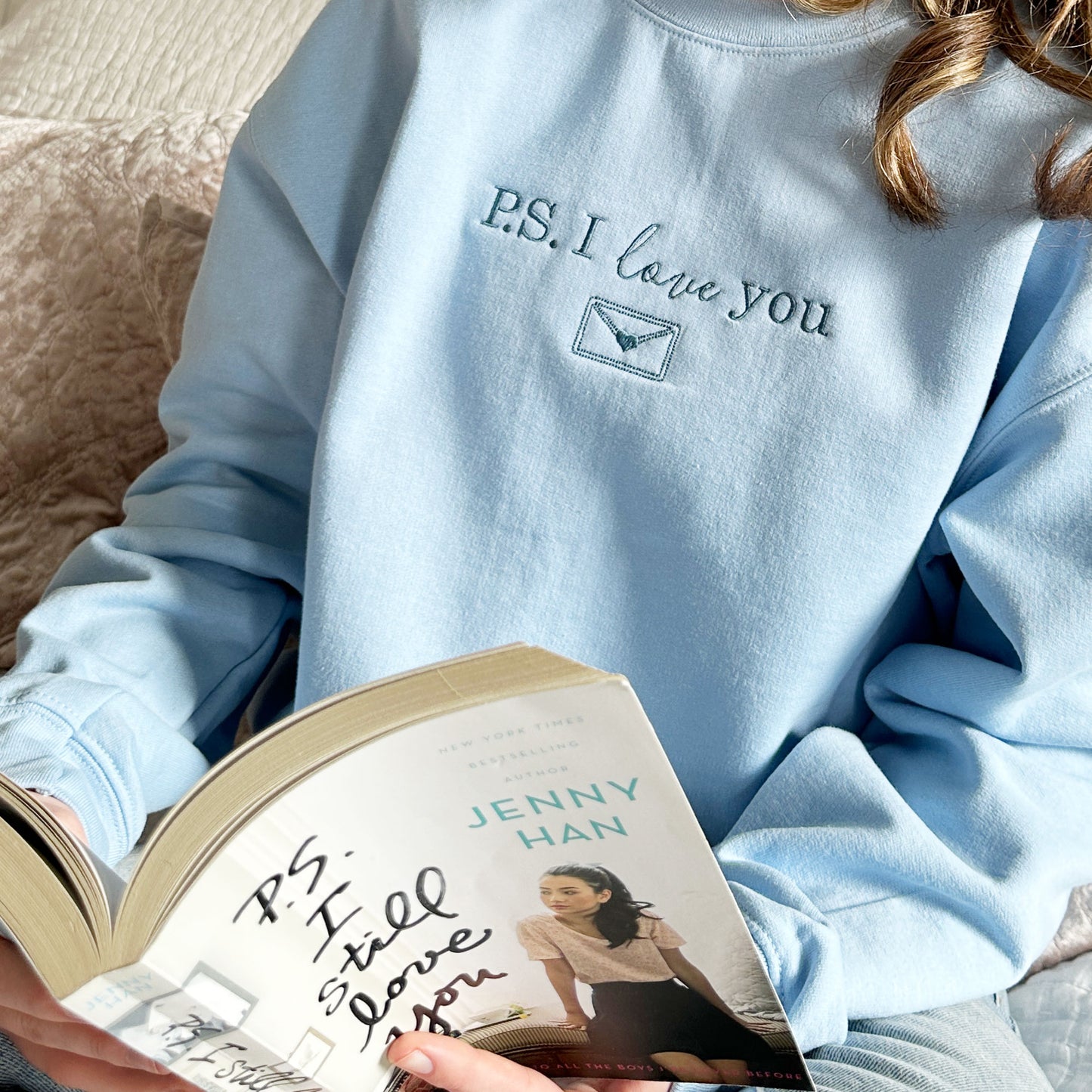 close up of a woman wearing a light blue crewneck sweatshirt with French blue embroidery that  says PS i love you with an envelope design underneath