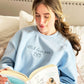 young women reading a book and modeling a light blue crewneck sweatshirt with PS I love you embroidery and an envelope design underneath the text