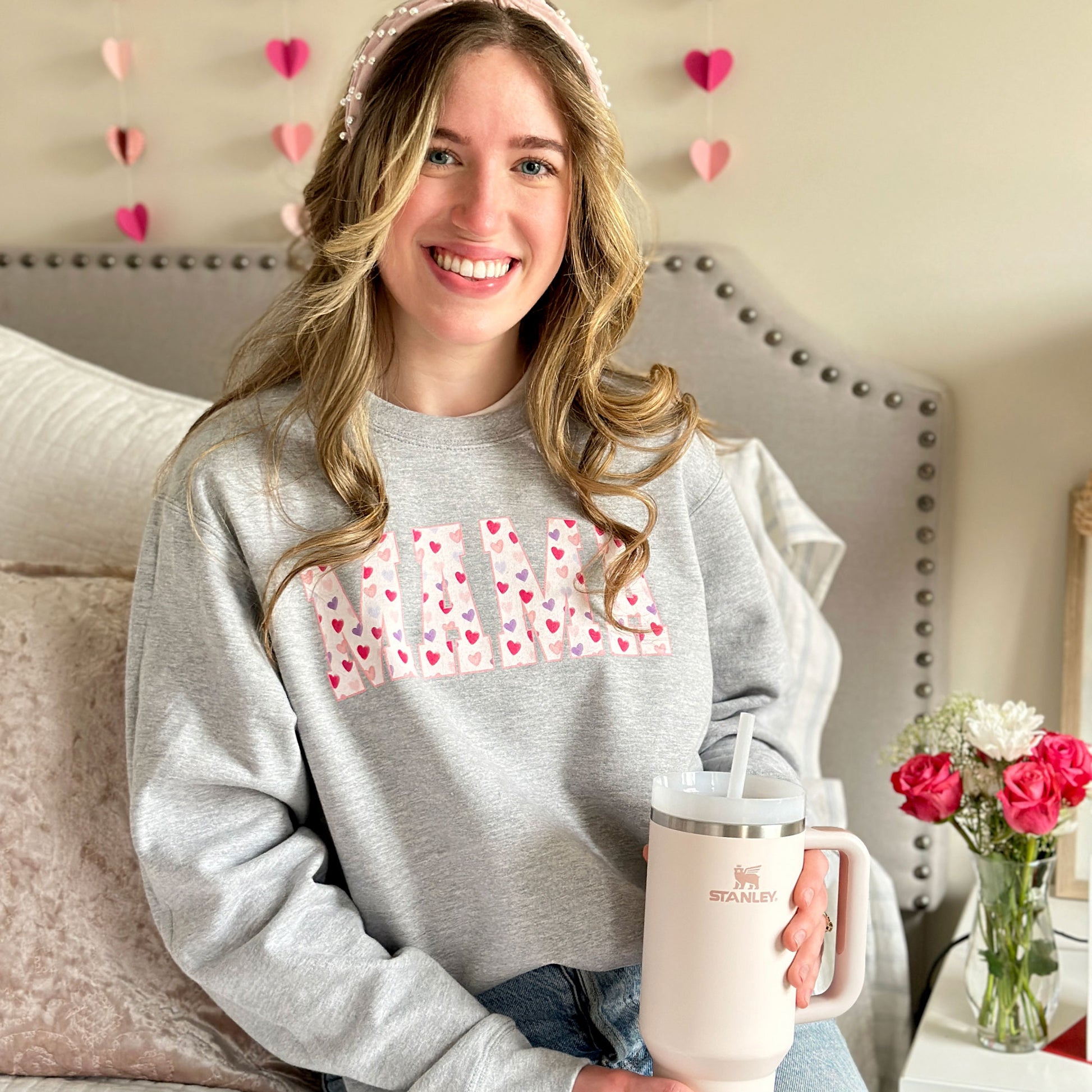 woman modeling an athletic heather crewneck sweatshirt with MAMA printed in a an athletic font filled with valentines hearts 