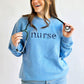 woman wearing a light blue crewneck sweatshirt with floral lowercase letters saying nurse in navy thread embroidered across the chest