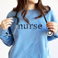 Close up of a woman wearing a light blue crewneck sweatshirt with lowercase floral letters that spell nurse in navy thread.