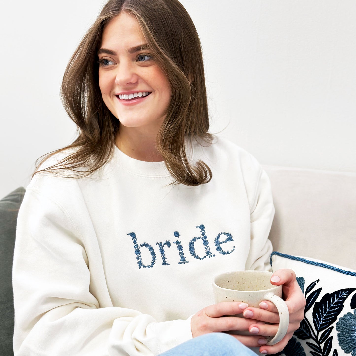Young woman drinking coffee on a coach modeling a white crewneck sweatshirt that says bride in lowercase blue floral lettering embroidered across the chest