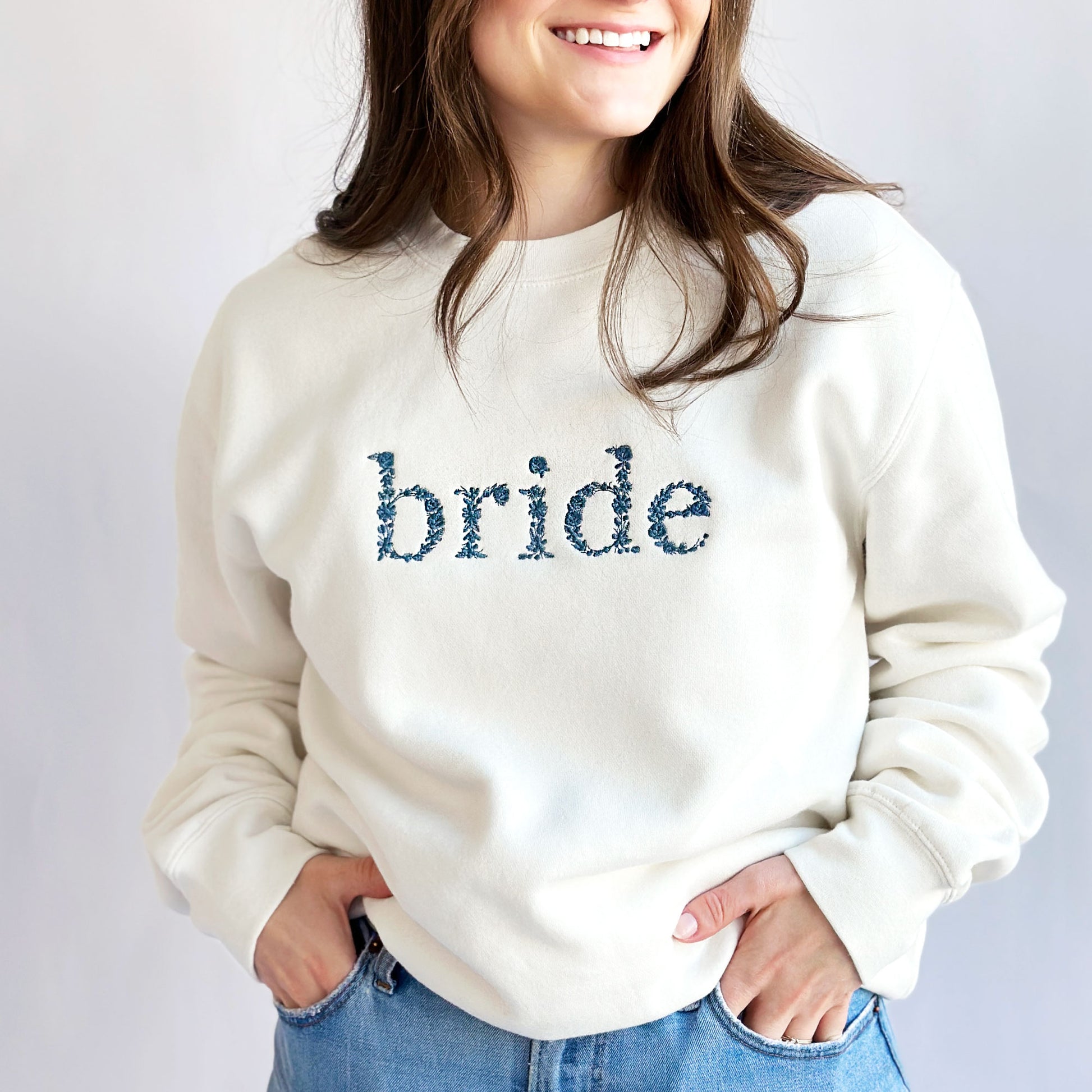 Young woman modeling a white crewneck sweatshirt with bride embroidered in floral lettering across the chest in French blue thread
