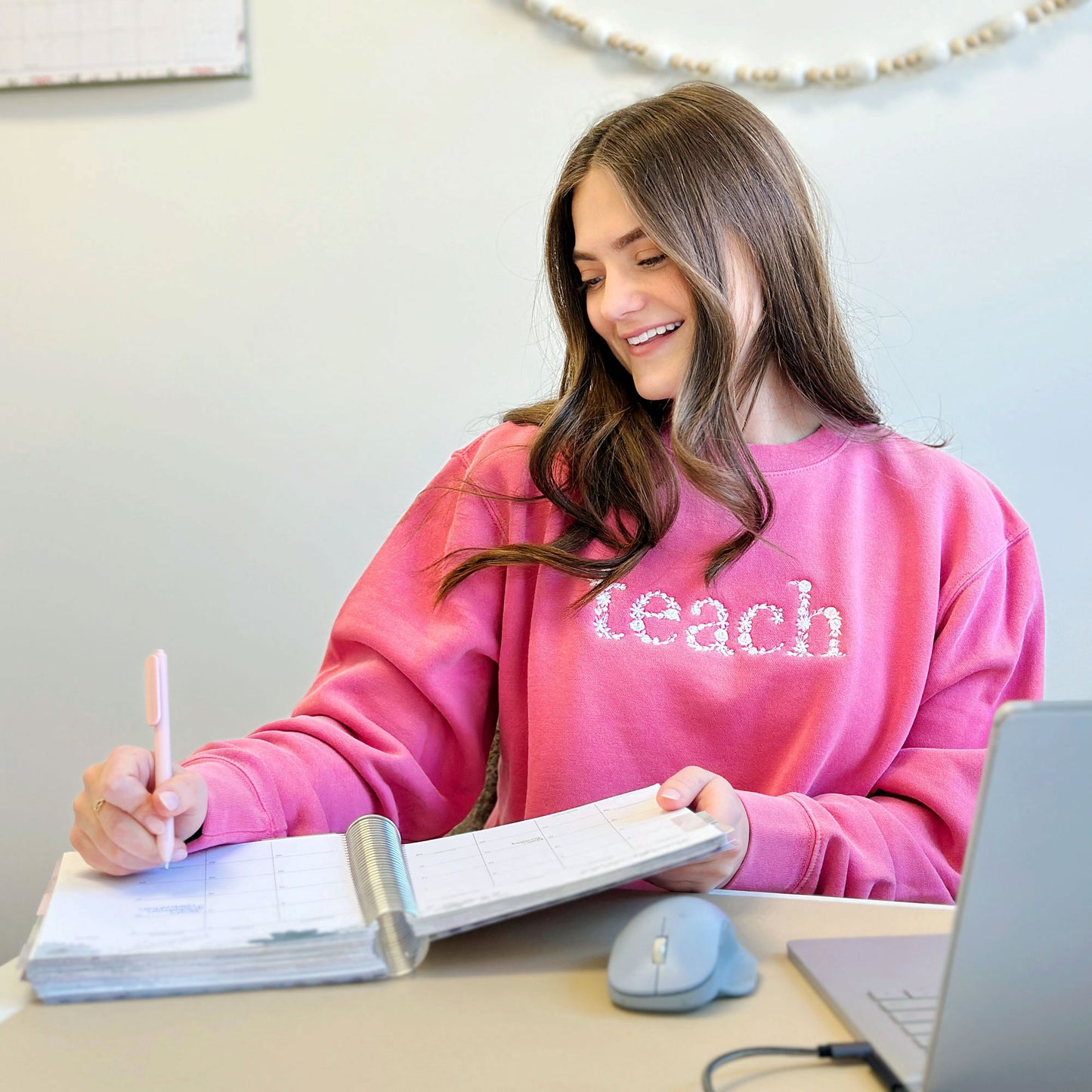 Woman at a desk wearing a pink crewneck sweatshirt with white floral letters teach embroidered across the chest