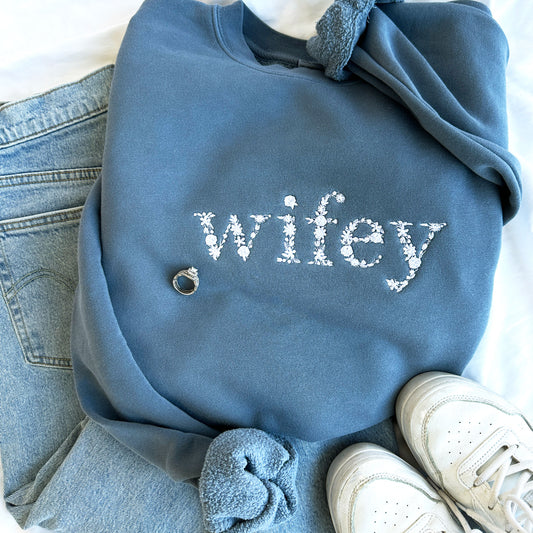 Flat lay of a slate blue crewneck sweatshirt with floral  lettering embroidered wifey design in white thread