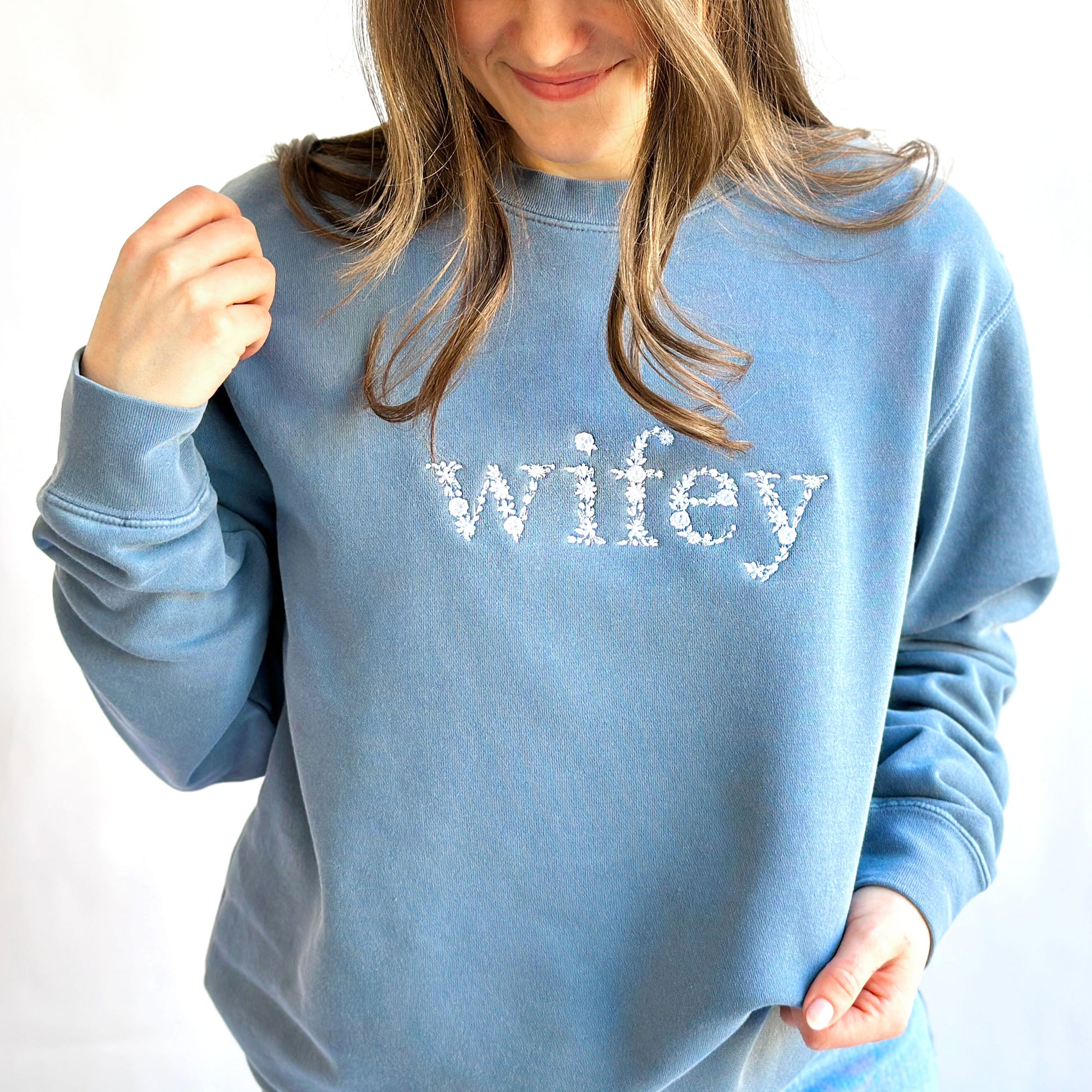 Young woman wearing a slate blue crewneck sweatshirt with widey embroidered in all lower case floral letters across the chest in white thread