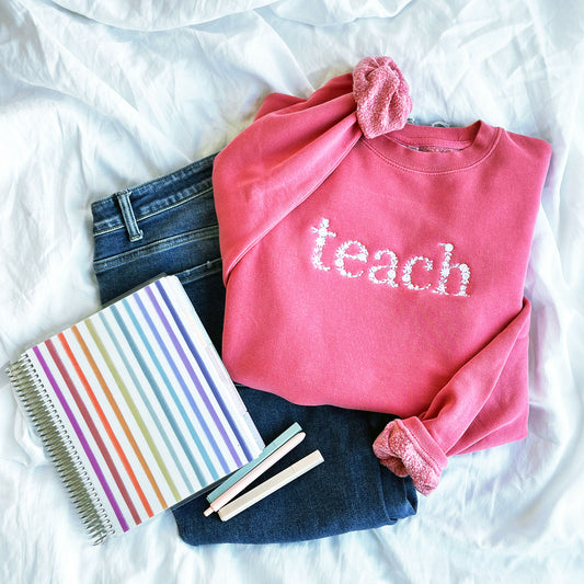 Flat lay of a pink crewneck sweatshirt with jeans, a notebook, and pens. The pink crewneck sweatshirt has float letters tach embroidered in lowercase letters across the chest in white thread.