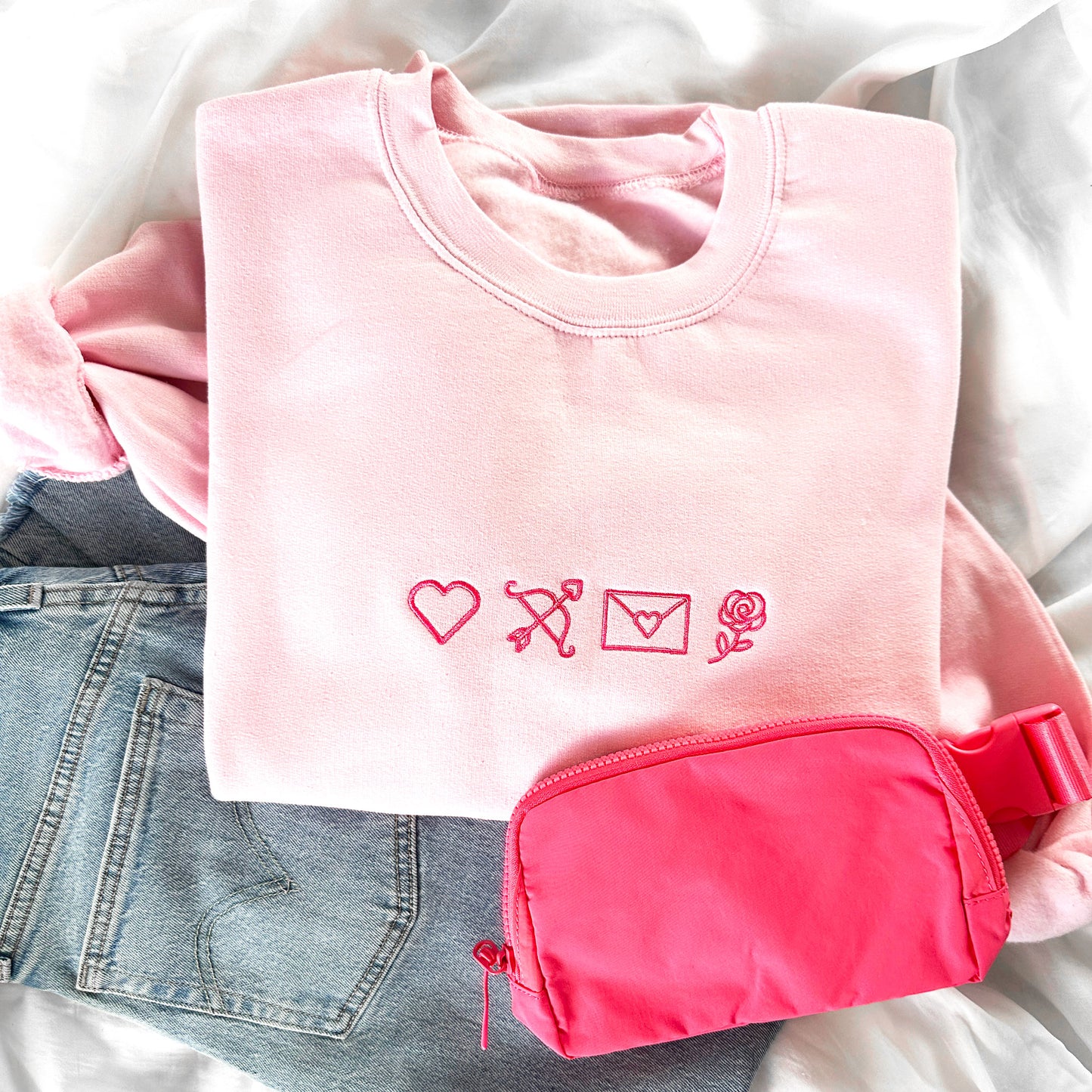 Styled flat lay of a light pink crewneck sweatshirt, jeans, and a pink fanny pack. on the center chest of the crewneck is 4 valentines icons embroidered in pink thread