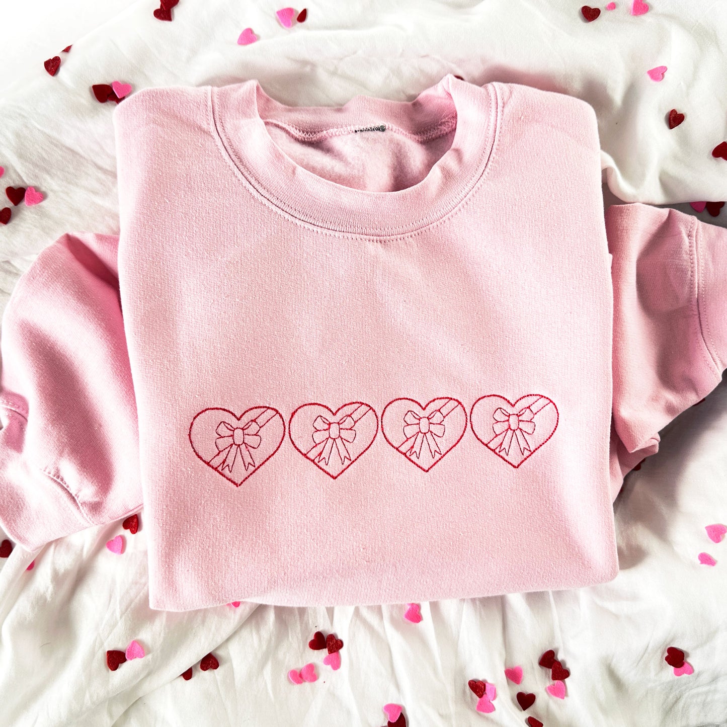 flat lay photo of a light pink crewneck sweatshirt with 4 stitched chocolates heart boxes with bows embroidered across the chest is raspberry thread