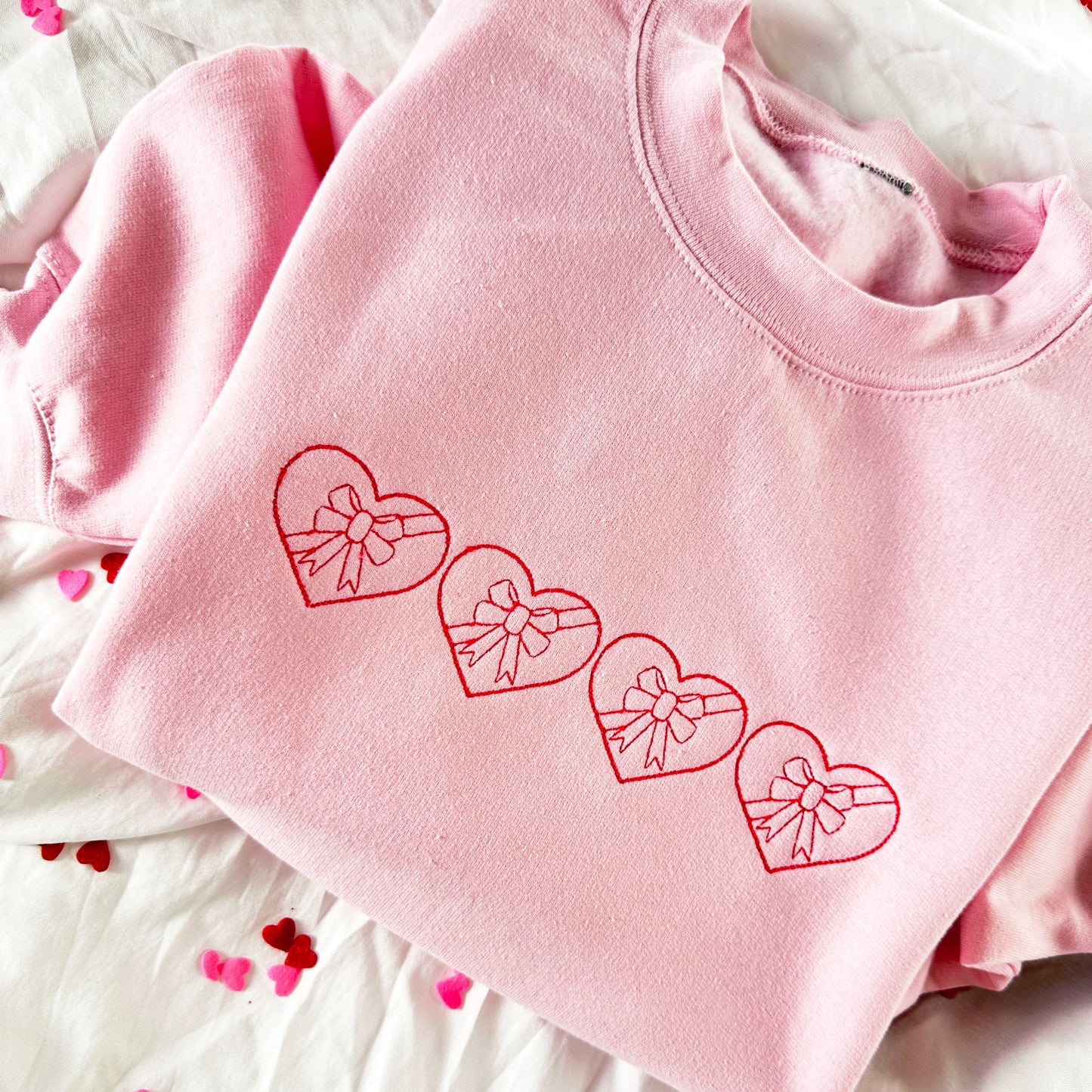 upclose of a light pink crewneck sweatshirt with 4 stitched hearts with bows in them across the chest for valentines day 