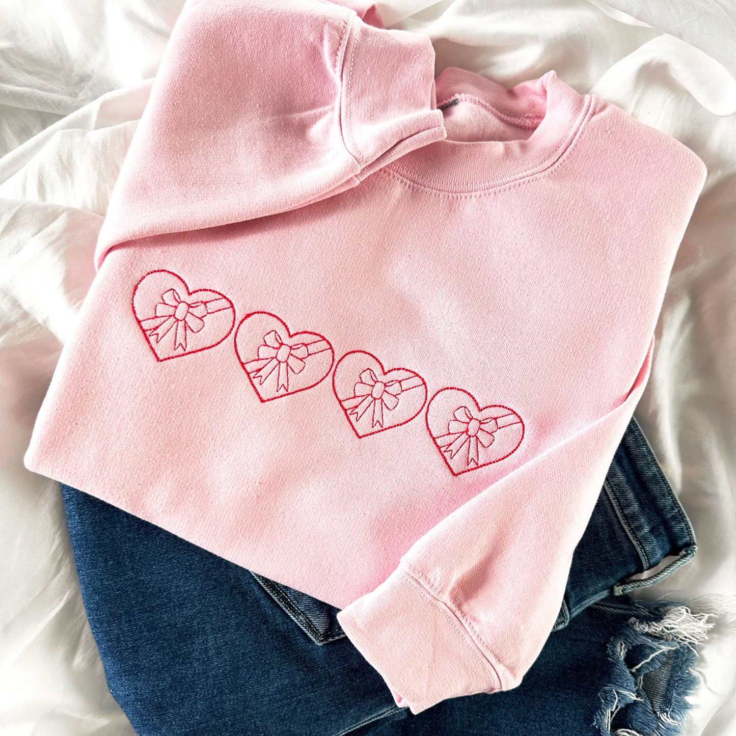 flay lay of a light pink crewneck sweatshirt and jeans. on the sweatshirt if 4 stitched valentines hearts with bows embroidered across the chest in raspberry thread
