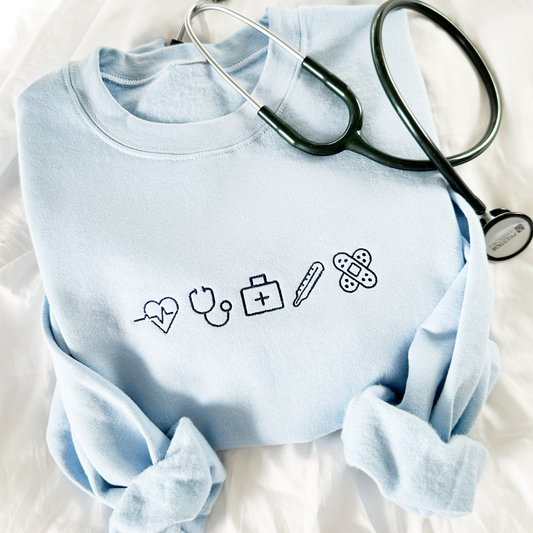 Flat lay of a light blue crewneck sweatshirt ith small nurse ions embroidered across the chest in navy thread