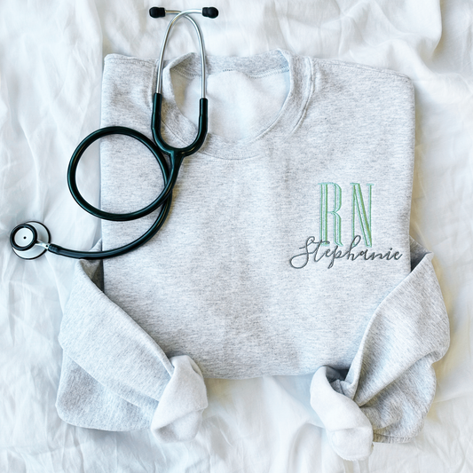 flat lay of an ash crewneck sweatshirt with embroidered nurse Rn and name design in silver sage and gray threads