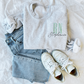 flat lay of an ash crewneck sweatshirt with embroidered nurse Rn and name design in silver sage and gray threads styled with jeans and sneakers