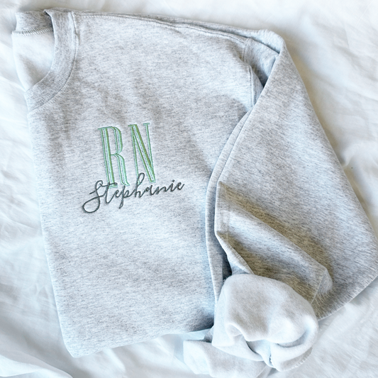 flat lay of an ash crewneck sweatshirt with embroidered nurse Rn and name design in silver sage and gray threads