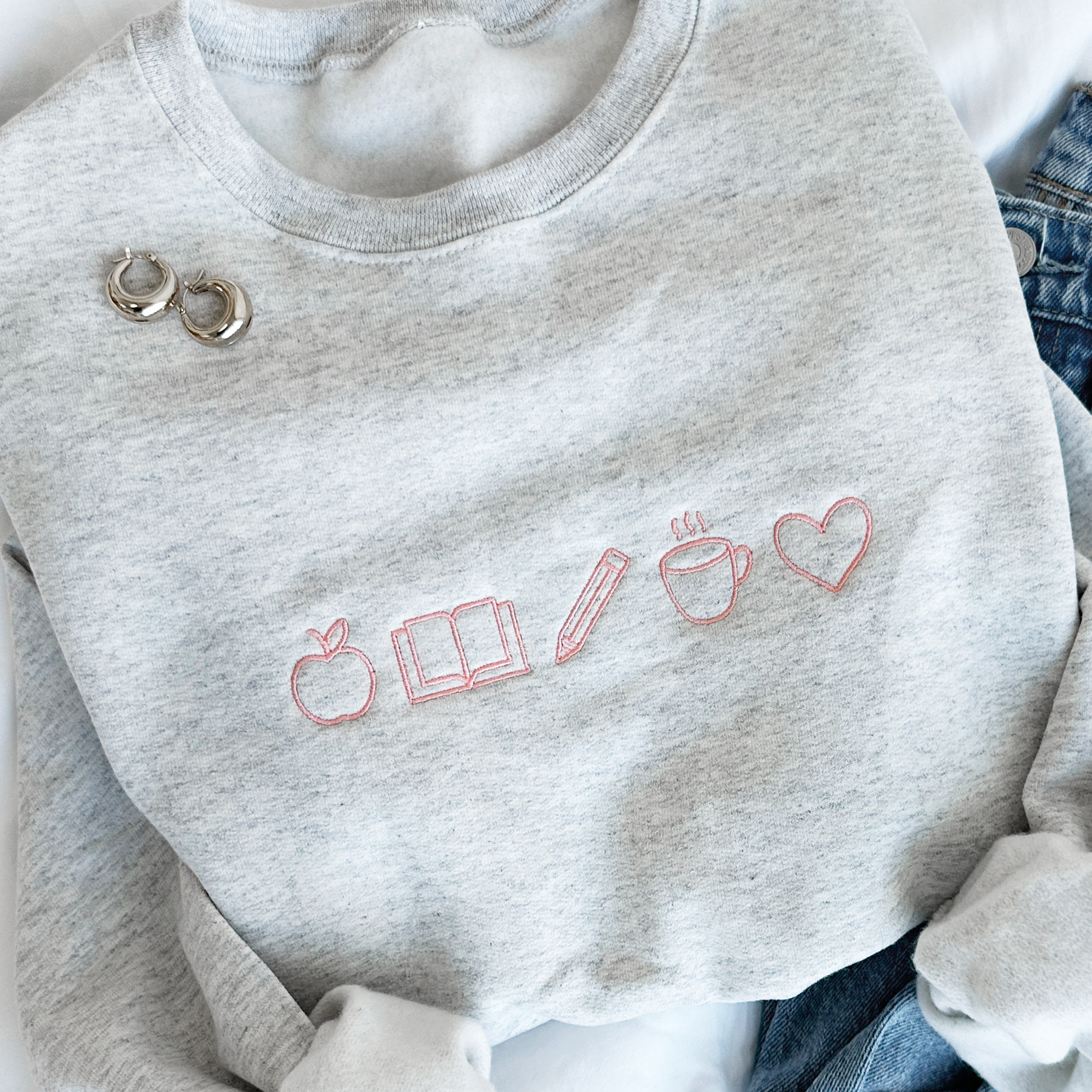 Flat lay photo of an ash crewneck sweatshirt with mini teacher icons embroidered across the chest in coral pink thread