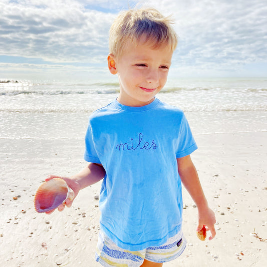 Little boy wearing a carolina blue t-shirt  with a stitch script custom name embriodery across the chest in a navy blue thread.