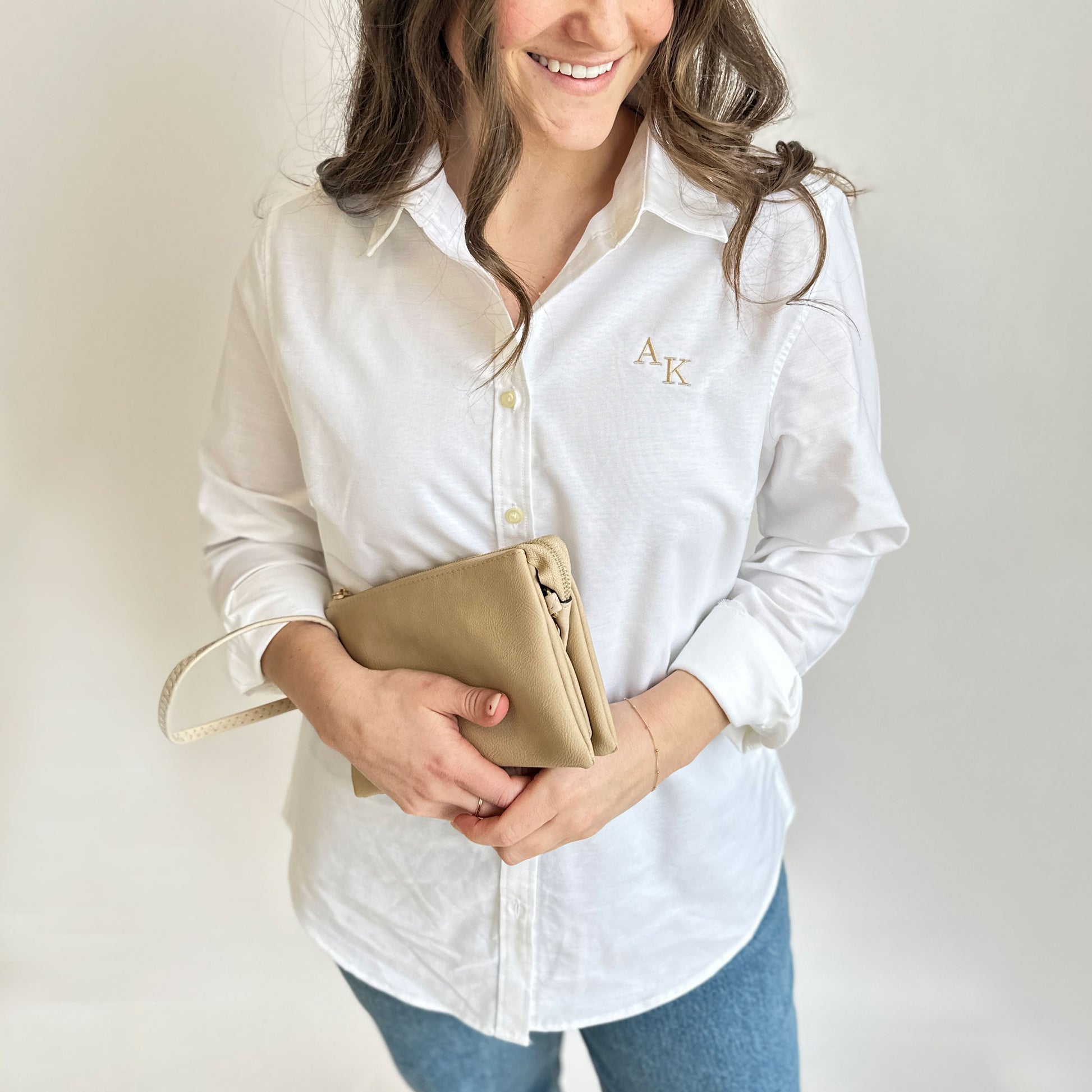young woman wearing a white oxford button down with mini AK embroidered on the left chest in camel thread, styled with a clutch purse