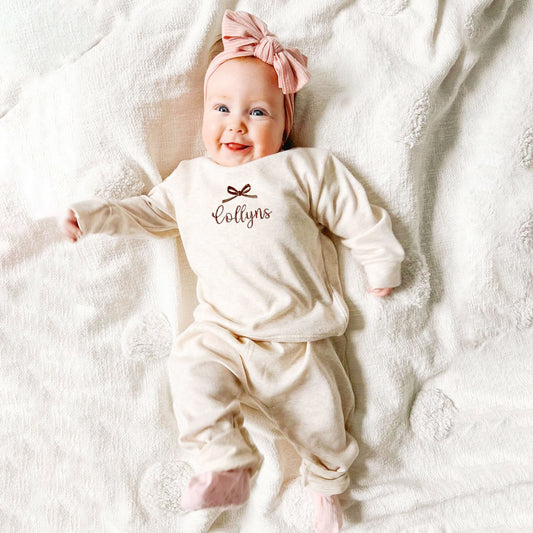 Baby girl wearing a 2 piece infant lounge set with custom bow and name embroidery on the center chest