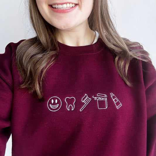 young woman close up selfie wearing a maroon crewneck sweatshirt with embroidered mini dental icons across the chest in white thread
