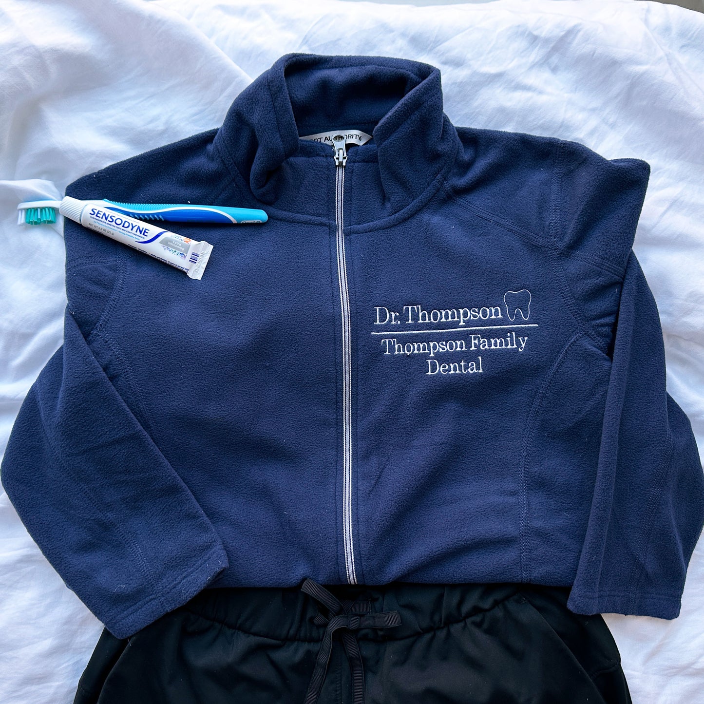 full zip navy fleece jacket with Dr, Thompson, Thompson family dental and mini outline tooth embroidered on the left chest on white thread styled with black scrubs and a toothbrush with toothpaste
